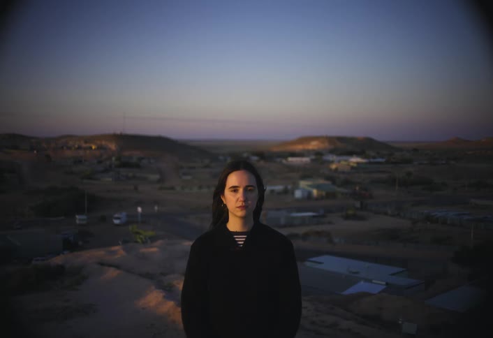 A dark haired woman wearing a black shirt stands on a hilltop looking straight at the camera with a sprawling dimly lit rural town visible below her under a dusk sky