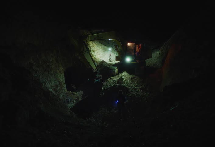 A worker walks toward a stationary excavator in a dark rocky excavation site