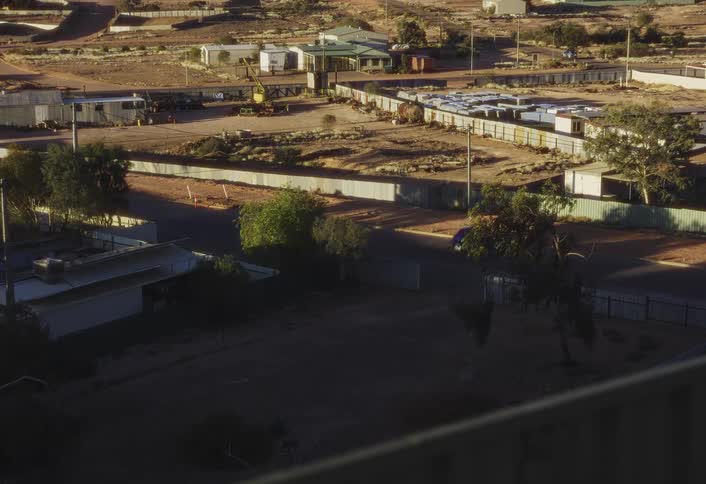 A high angle wide view shows a blue car driving through a dusty remote mining or industrial town with numerous small buildings construction supplies and open red dirt yards