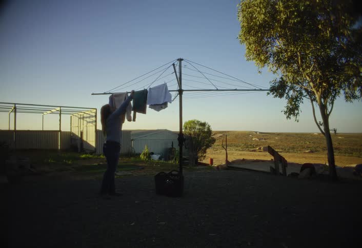 A young woman is hanging clothes on a rotary clothesline in the yard of a remote dusty rural property with farm buildings and a large dry field visible under a bright sky