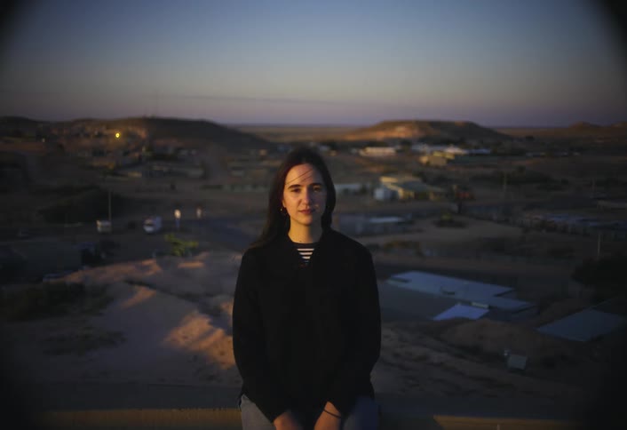 A young woman stands atop a hill at sunset dressed in a black longsleeve tshirt with a small town sprawling behind her