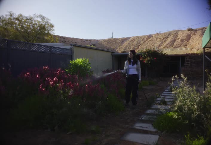 A woman waters flowers in a sunny backyard garden with desert hills behind