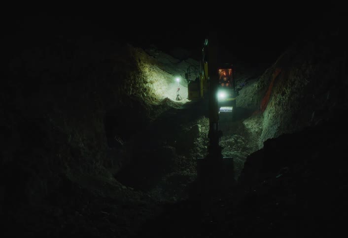 An excavator digging a hole outdoors at night with the area illuminated by lights