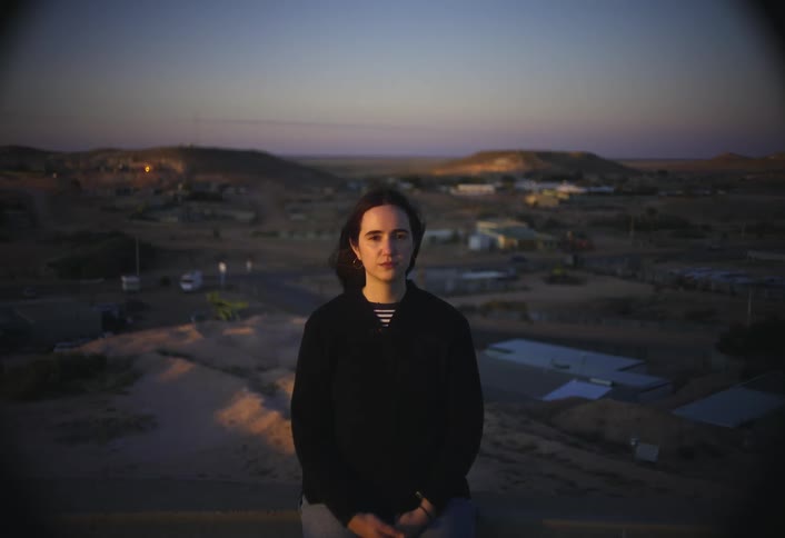A young woman wearing a black shirt stands outdoors on a hill as a light wind blows her hair at sunset with a small town in the background