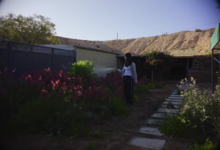 A young woman in her backyard sprays water from a hose onto her garden plants at midday