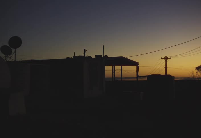 A wide dark silhouette of a small building with satellite dishes and a telephone pole is set against a vibrant orange and blue sky at sunset or dawn in a remote location