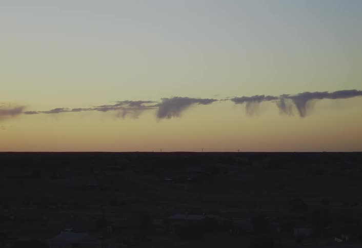 A long wide view of a dark horizon shows thin elongated clouds with rain streaks against a pale yellow and orange sunset sky over a flat undeveloped landscape