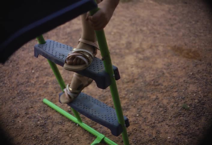 A closeup shot focuses on a young childs legs and feet as they carefully climb a small playground ladder with perforated gray steps the child wearing tan sandals and shorts slowly navigates the brightly colored green rungs against a backdrop of dirt