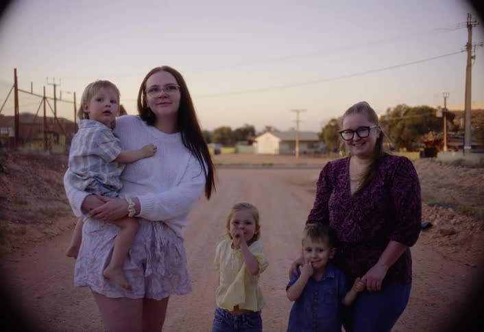 Two women and three children are standing on a dusty dirt road in a remote town smiling for the camera as the woman on the left holds a young boy