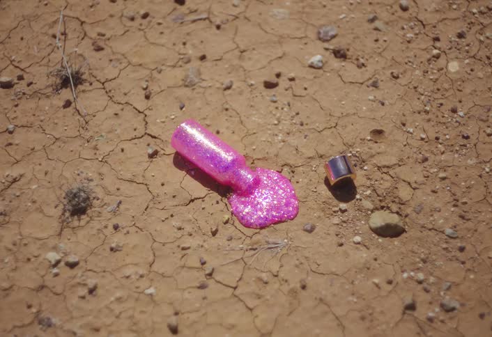 This closeup shot features a small bottle of pink glitter nail polish spilled and lying on dry cracked dirtthe polish is glistening brightly in the sun and the metal cap of the bottle is resting nearby