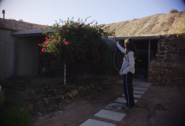 A young woman in the backyard of her house uses a hose to water plants on a sunny day
