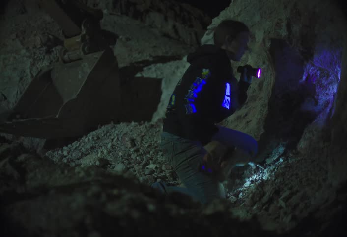 A woman kneels on a pile of dirt in a dark trench at night wearing a black hoodie and jeans while using a purple ultraviolet flashlight to examine a light colored dirt and rock wall next to a large excavator bucket