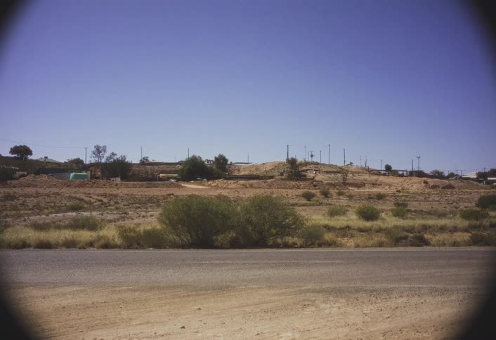 A roadside cuts through a desert landscape with distant hills under a clear blue sky