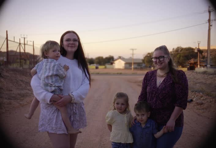 Two women and three children are standing on a dusty dirt road in a remote town smiling for the camera as the woman on the left holds a young boy in the soft sunset light