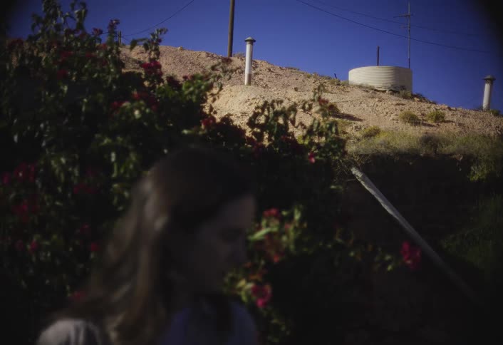 A slightly blurred woman stands in the foreground next to dark green bushes and red flowers while a dry hill with a large white water tank is visible against a bright blue sky
