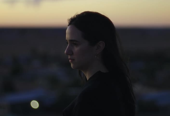 A profile close view shows a womans dark silhouette against a pale orange sunset sky with a blurred remote desert town visible in the background