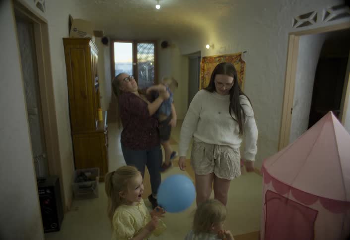 Mother puts down her son while another woman blows a kiss to children playing indoors with a castle play set