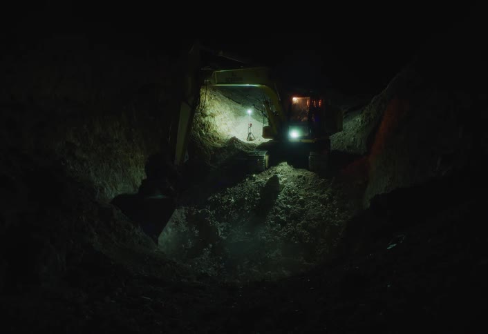 A yellow excavator digs and dumps dirt out of a deep dark trench at night illuminated by the bright work lights on the machine and casting strong shadows on the earthen walls