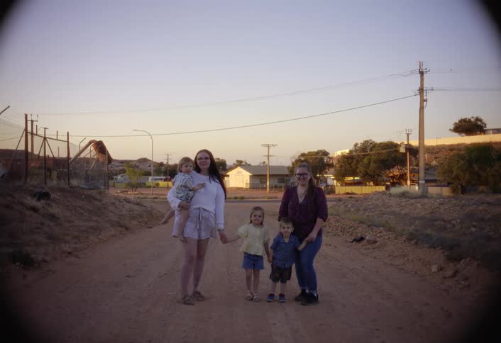 Two adult women one holding a baby and one standing with two young boys pose and look directly at the camera on a wide unpaved dirt road in a possibly rural or outback setting with utility poles and fenced structures against a pale sky