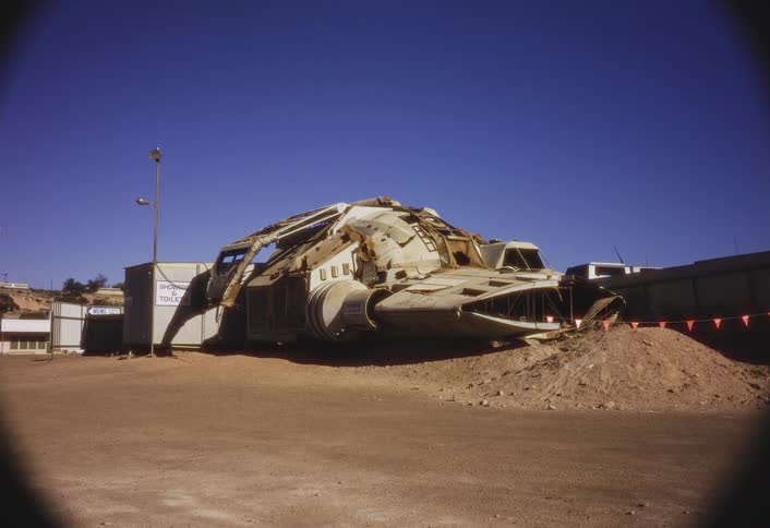A damaged futuristic white construction vehicle is half buried in a brown dirt mound in a dusty outdoor lot next to small buildings under a clear blue sky