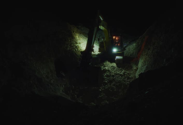 A deep dark trench is carved into the earth at night illuminated by the bright work lights and cab of a yellow excavator operating in the tight space