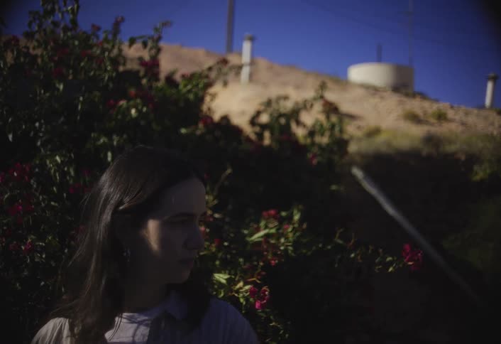 A young woman with long dark hair stands in the foreground partially framed by a dark green bush with red flowers while the background reveals a sunny dry hillside topped with a large round water tank and ventilation pipes against a clear blue sky