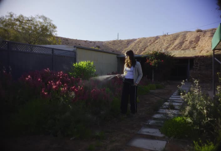 A young woman wearing a white shirt and dark pants stands in a dry outdoor garden watering a large bush of red flowers in front of small rural buildings under a bright blue sky