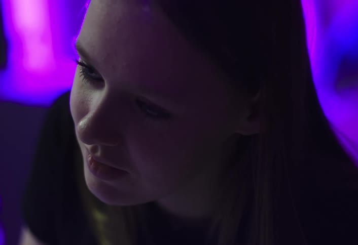 A young girl sits inside a room illuminated by blue and purple led lights appearing to be engaged in a conversation with someone