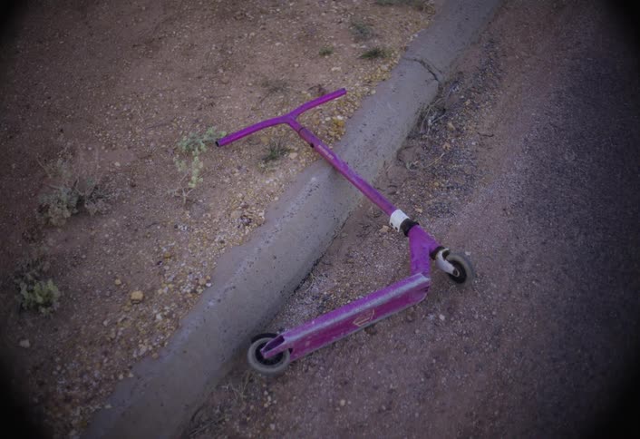 A bright pink scooter is lying abandoned on its side against a gray concrete curb next to a brown dirt road seen from a high angle