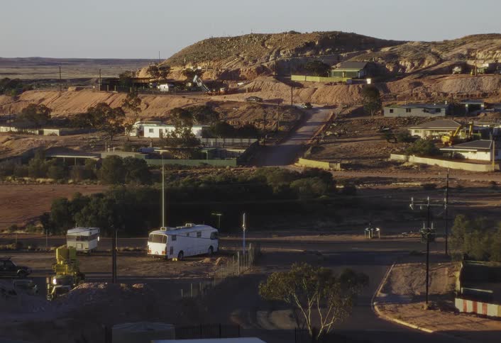 A sunlit desert hillside town with scattered low buildings winding dirt roads and a few parked and driving vehicles near rocky terrain