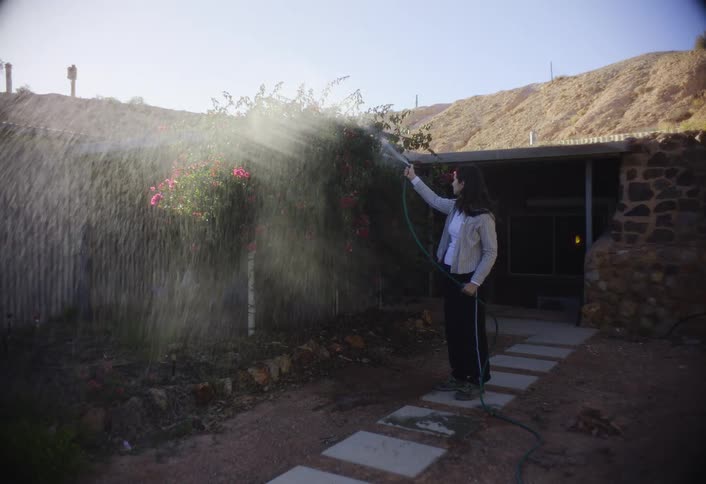 Woman standing outside her house waters a flowering bush during the day