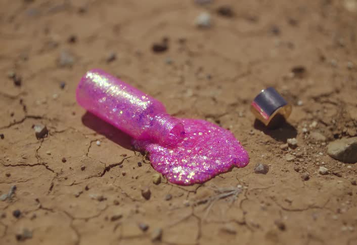 A bottle of bright pink glitter nail polish is spilled and running onto the cracked brown dirt of a dry ground with its black and gold cap lying separately in the sunlight