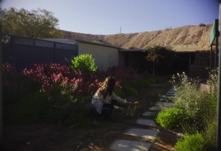A woman kneels in a garden petting an orange cat on a sunny day