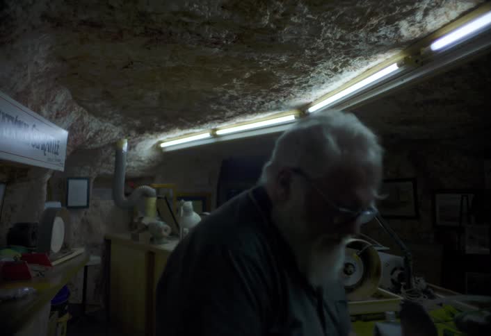An elderly man removes his face goggles after finishing work inside a workshop then switches off the light as he walks out
