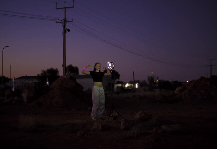 A teenage girl performs in front of her cell phone in a dirt lot