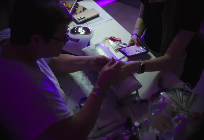 A female nail technician wearing eyeglasses is trimming the cuticles of a customers hand with clippers while the customer holds up a phone displaying nail color videos under bright pink ambient light in the salon