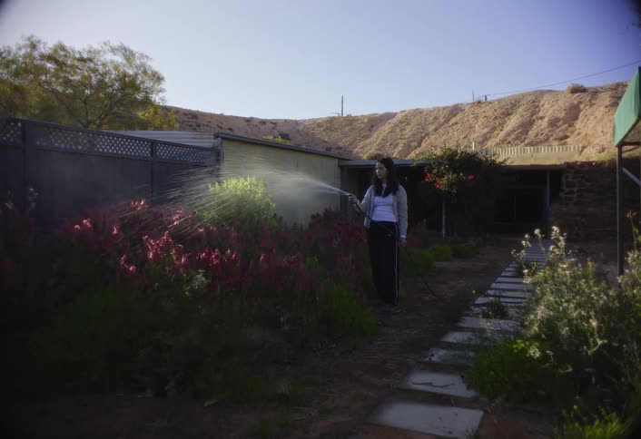 A woman watering plants in a dry backyard below a bare hill