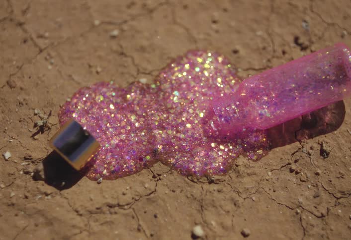 A container falls onto sandy soil spilling its contents a pink lip gloss with glitter at midday