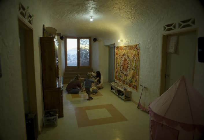 A mother sits on the floor in her living room with a friend while her kids play around the room at midday