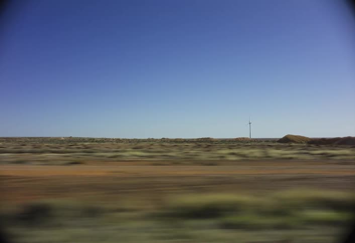 A low wide view from a moving vehicle shows a brown dirt road and sparse green scrubland with piles of earth and a small white wind vane visible on the flat horizon under a clear blue sky