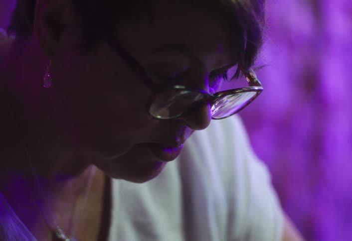 A very close view shows the side profile of a woman wearing eyeglasses and a white shirt talking and looking down intensely under dramatic pink and purple light