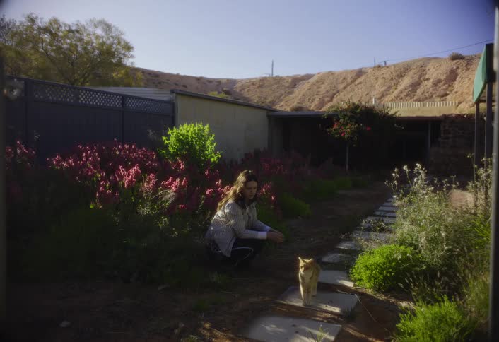 A woman pets a cat in a garden near a path with desert hills behind