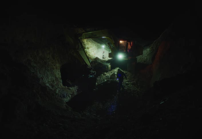 In a excavation pit a bright work light illuminates a yellow excavator positioned between high dirt walls while a worker wearing dark clothing briefly enters the frame in the foreground using a small blue light to examine the ground
