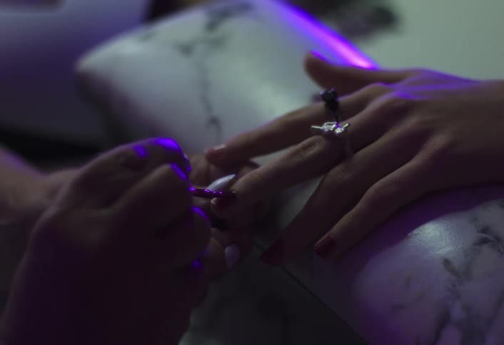A closeup shot under purple light shows a manicurist carefully applying polish to a customers fingernails the customer is wearing a distinctive ring and resting her hands on a marbled arm cushion during the beauty treatment