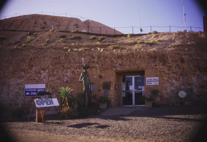 The entrance to a building carved into a reddishbrown arid hillside which is topped with lowlying green shrubs and a security fence featuring a central wooden statue glass doors and an open sign