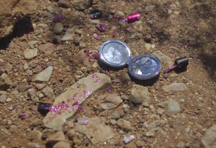 A broken pocket mirror lies open on rocky ground beside spilled pink glitter and nail polish