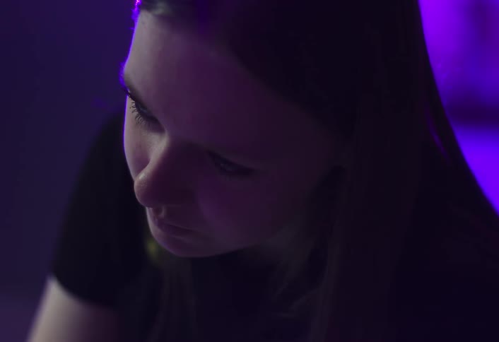 This is a closeup shot of a young womans face illuminated by a strong purple light from the side or backgroundshe is looking down but then slowly looks up and offers a slight smile before looking down again