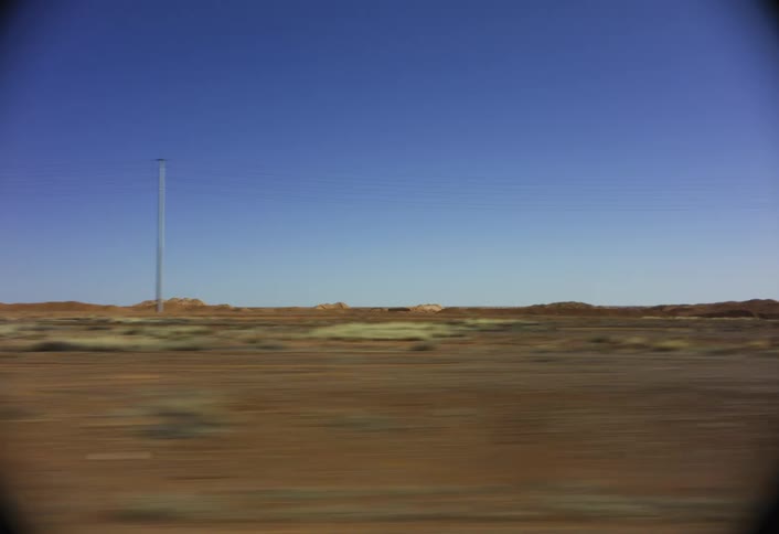 Captured from a side window of a vehicle in motion the shot sweeps across a flat redbrown arid plain beneath a bright blue sky with utility pole lowlying rock formations and manmade earth mounds