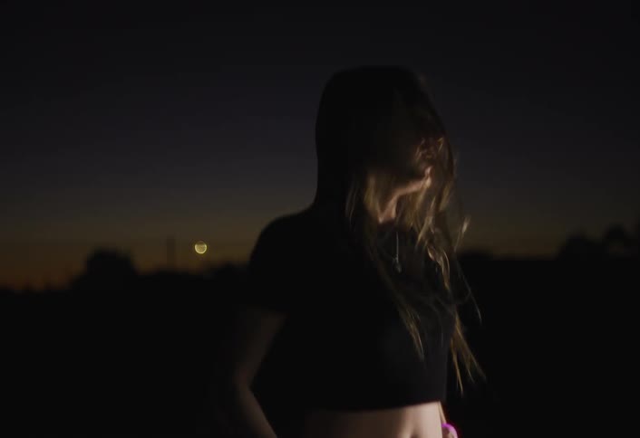 A young woman with long hair and a bare midriff is dancing energetically at dusk wearing pink and purple glow bracelets that illuminate her arms against a dark sky