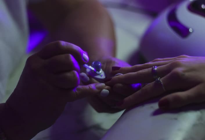 A manicurist is giving a woman a manicure in a close view applying clear coat polish to the red painted fingernails under a dramatic purple ultraviolet light